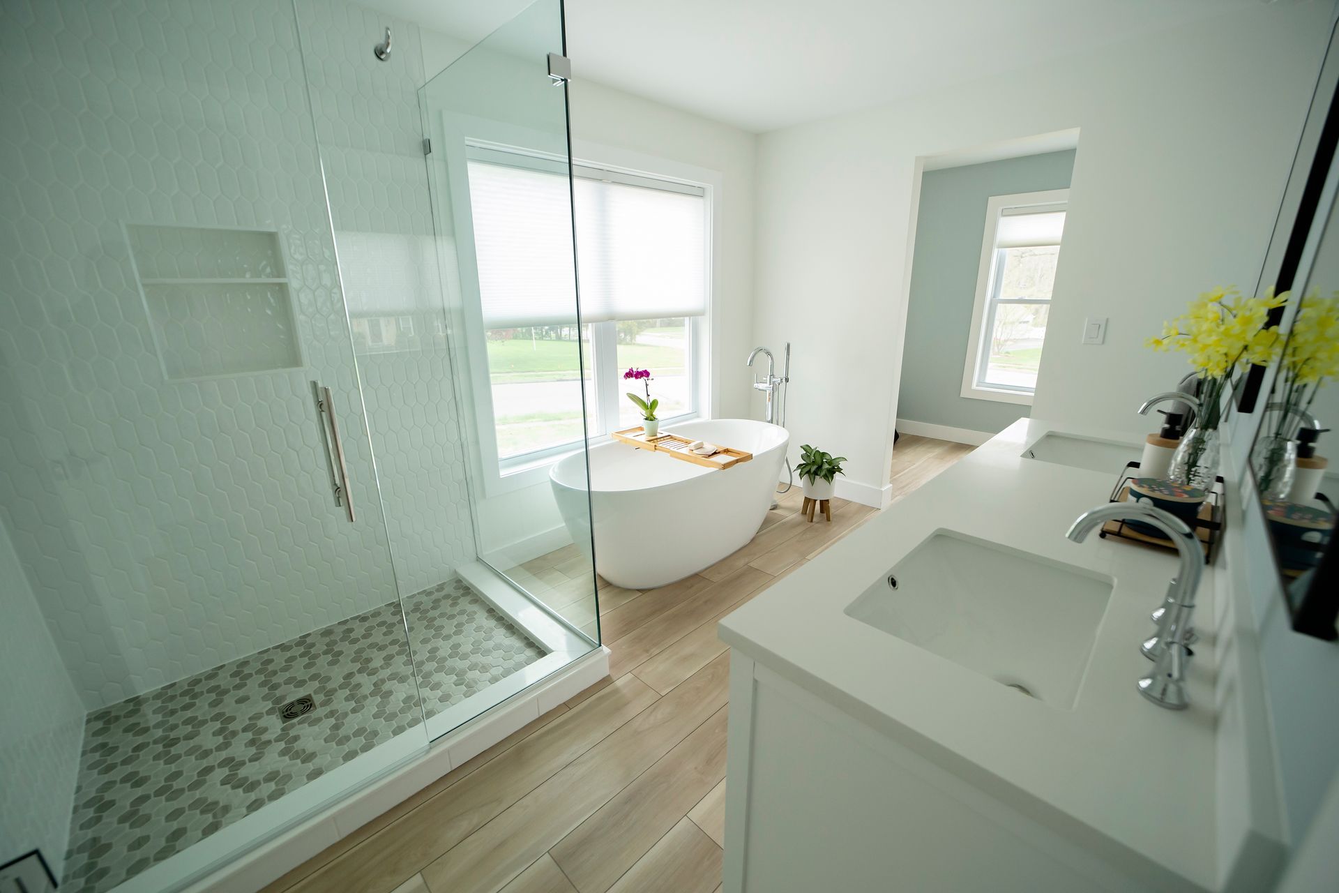 Bright modern bathroom with glass shower, freestanding tub, vanity, and large window.