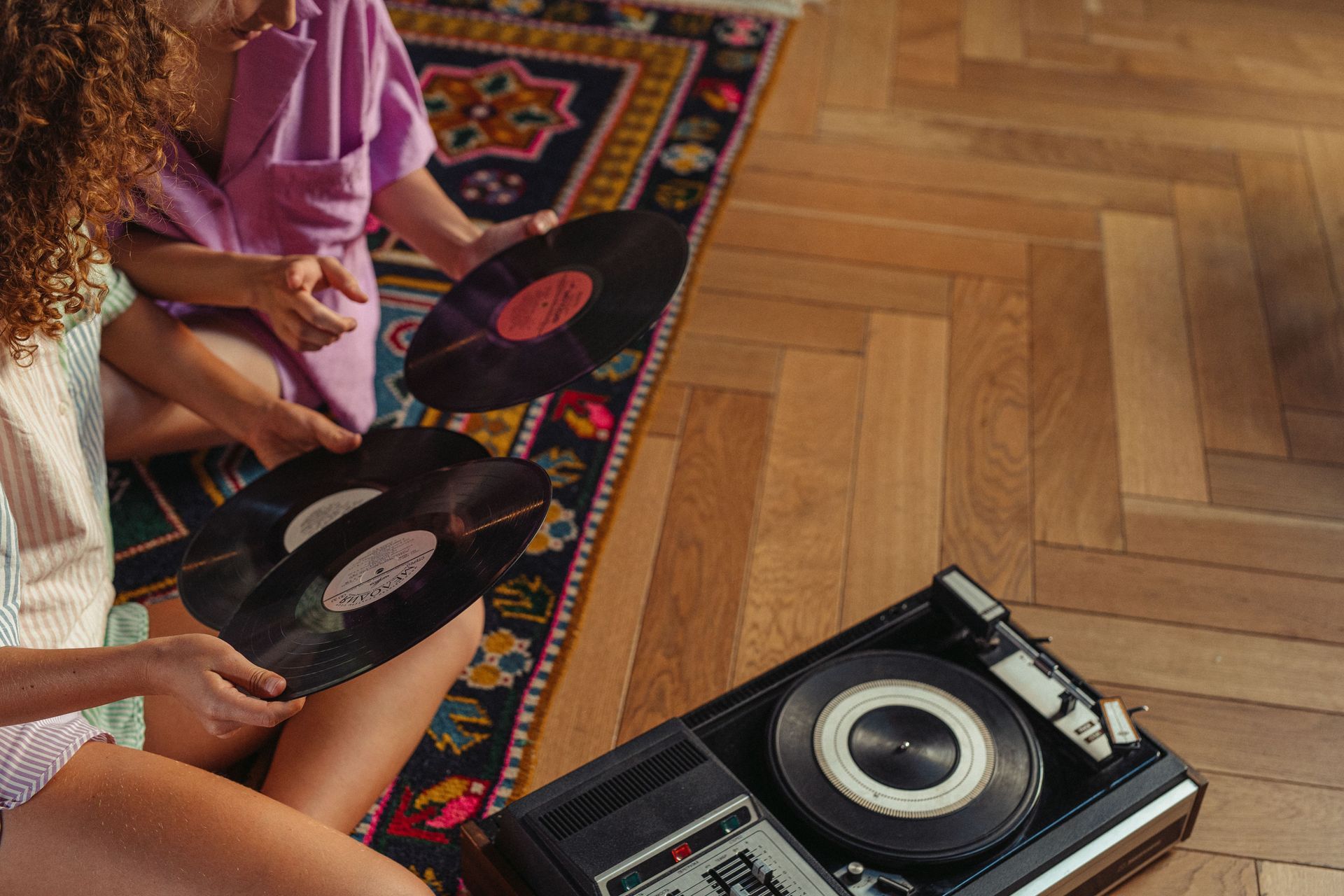 People holding vinyl records beside a turntable on a patterned rug and wooden floor
