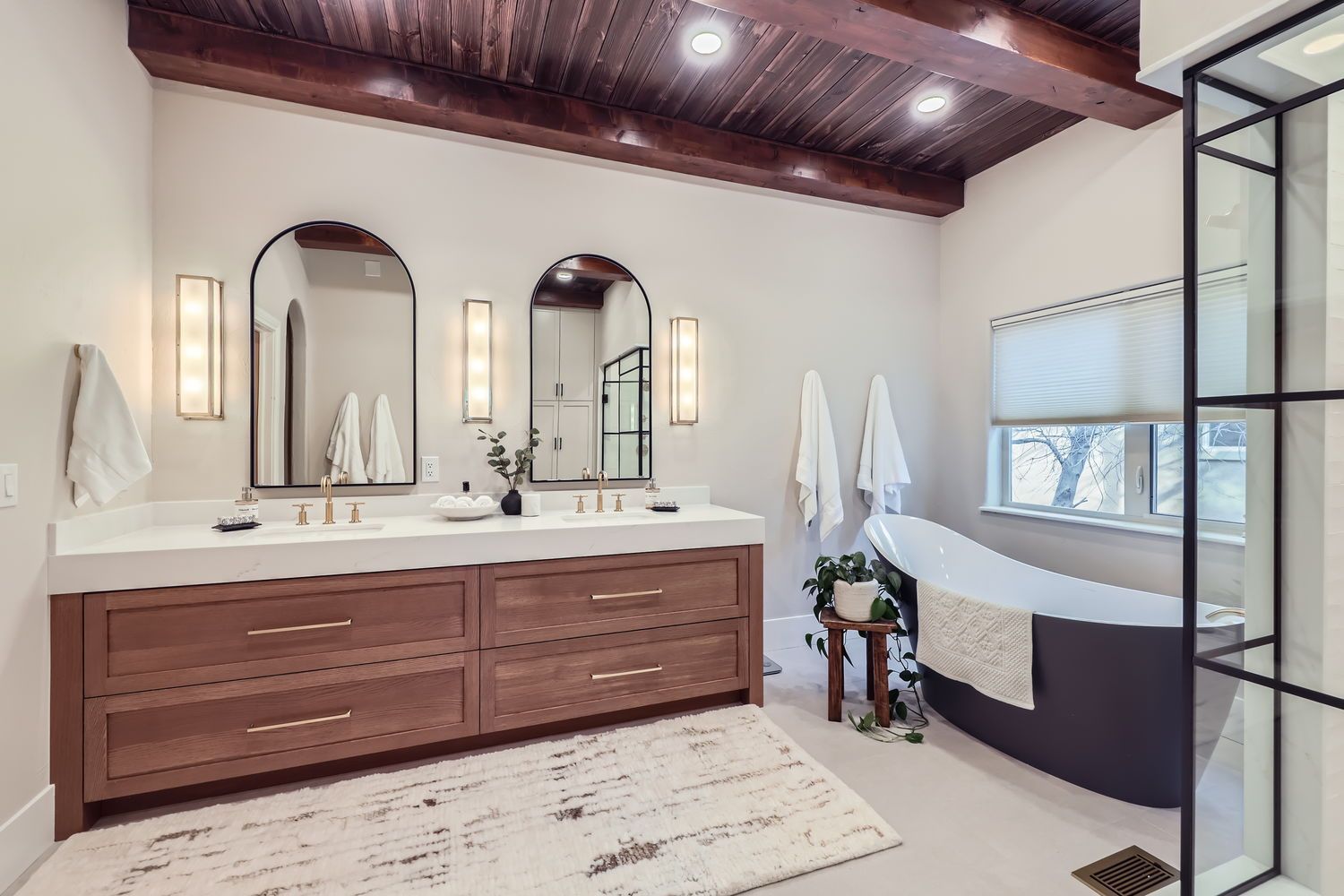 Modern bathroom with wood ceiling, double vanity, arched mirrors, and freestanding tub.
