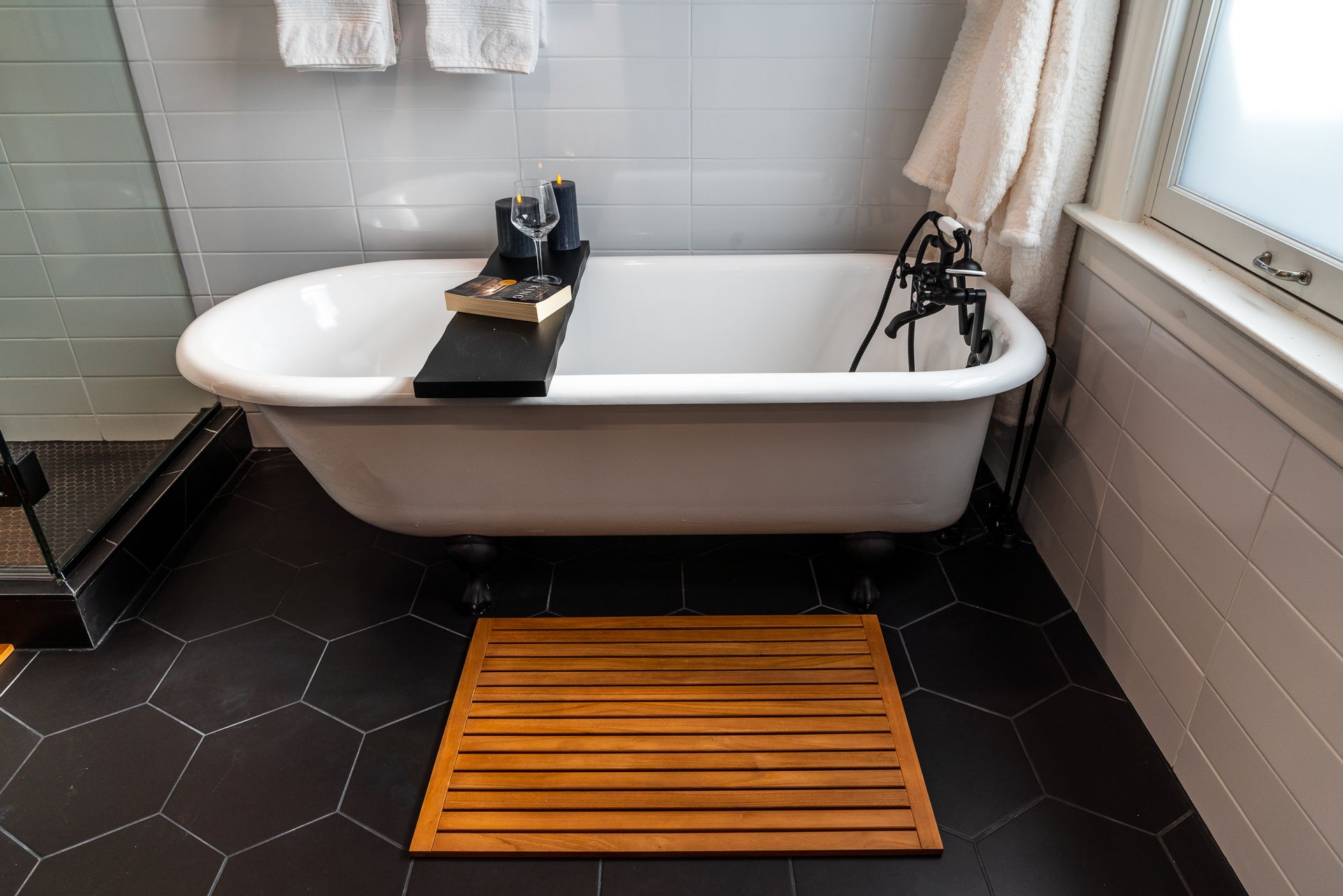 Clawfoot bathtub with wooden bath mat and black hexagonal tile floor in bathroom setting.
