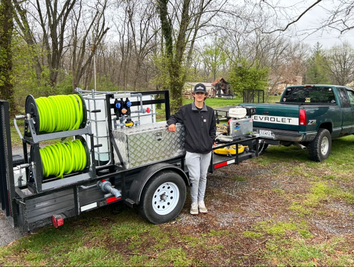 A young person stands by a trailer with pressure washing equipment, attached to a green pickup truck, outdoors.