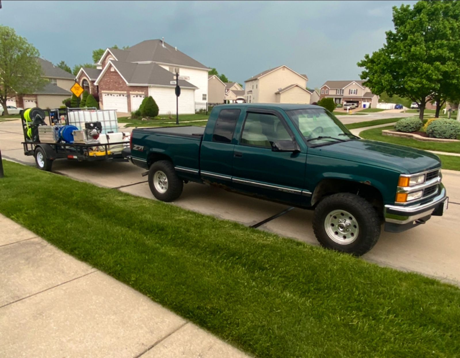 Green pickup truck towing a trailer with pressure washing equipment parked on a residential street.