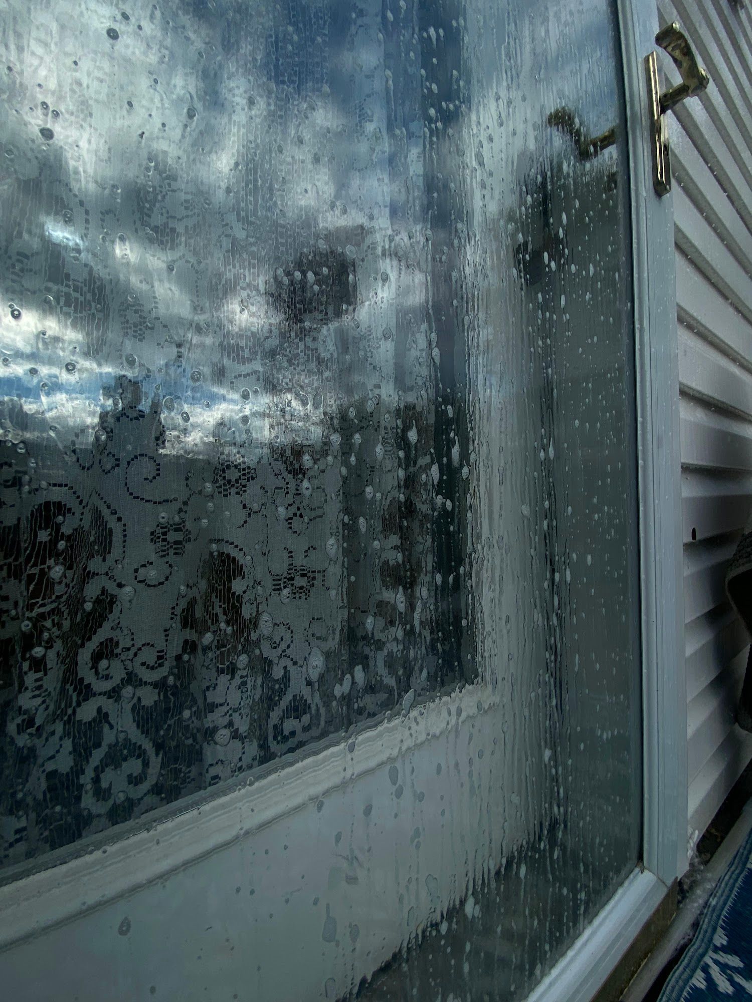 Window with water droplets obscuring the view of a cloudy sky and blurred outdoor landscape.