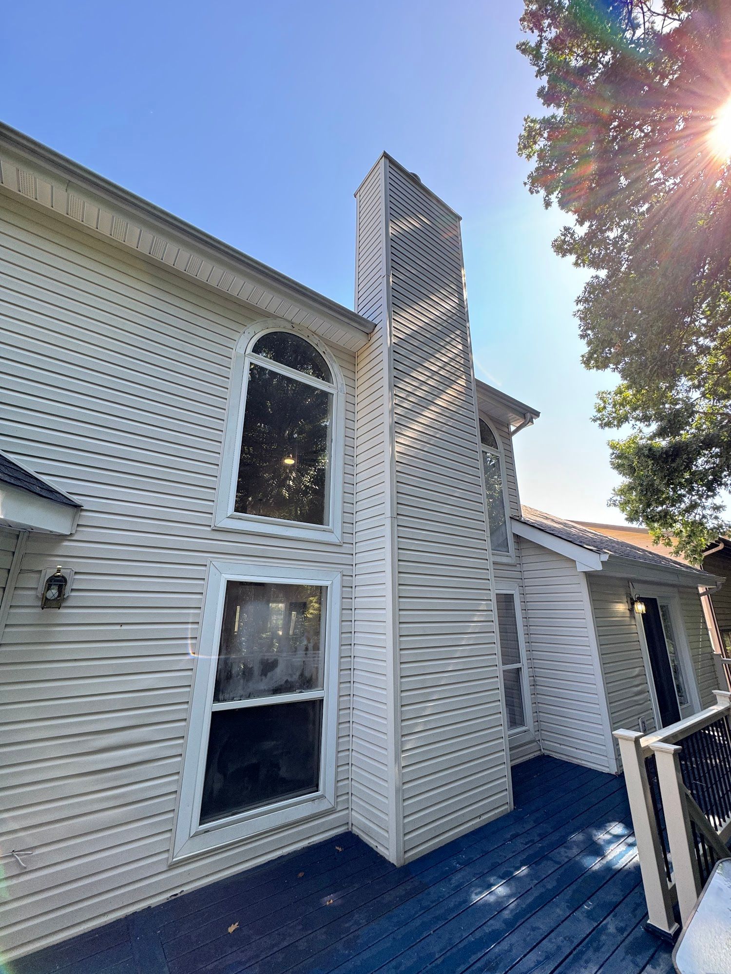 Exterior of a two-story house with a tall, decorative chimney. Blue deck, white siding, and bright sun.