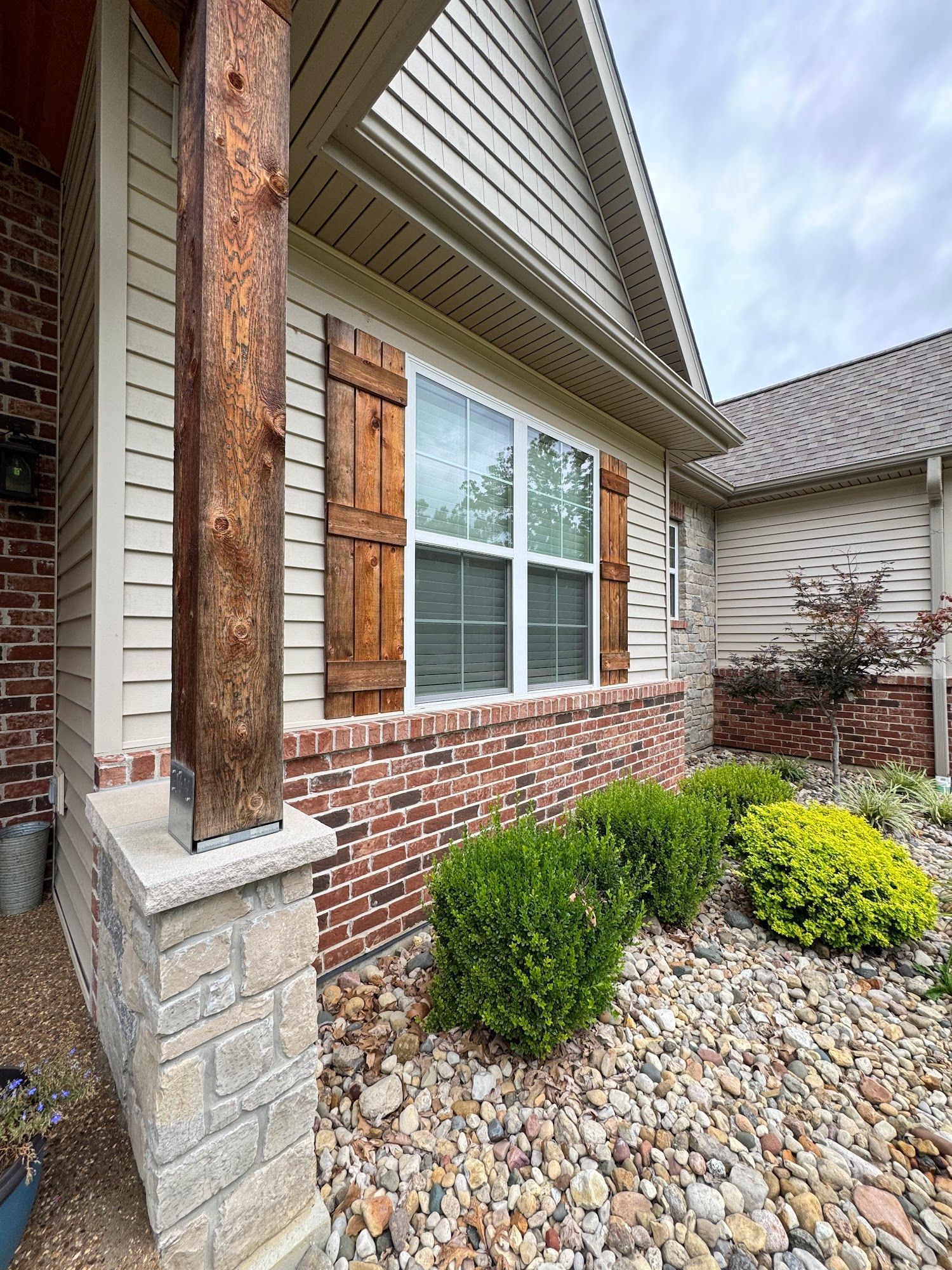 Exterior of a house with light siding, brick, and wooden shutters; the landscaping includes bushes and stones.