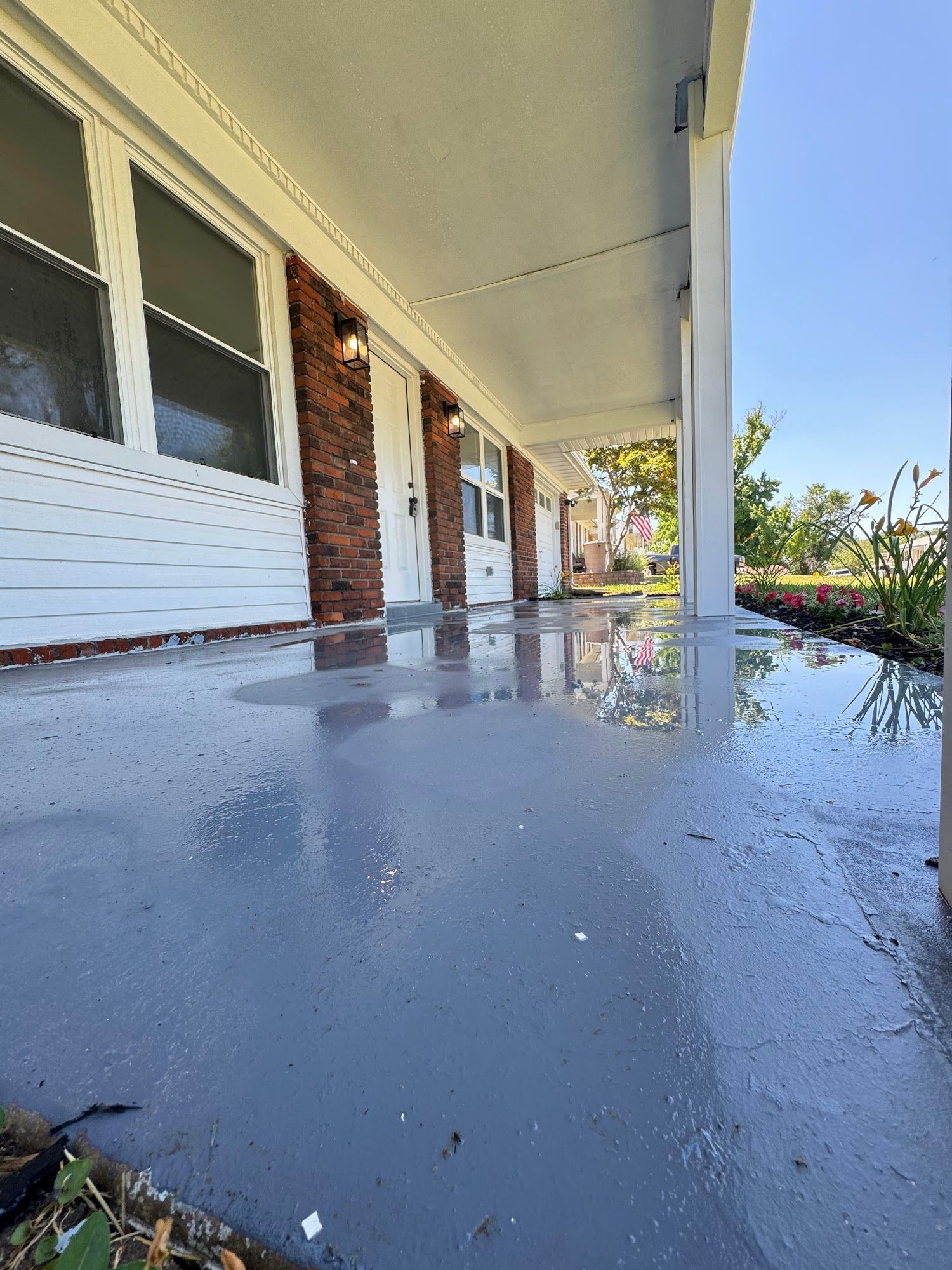 A gray painted porch reflects the sky. Brick, white siding, windows, and a column support the roof.