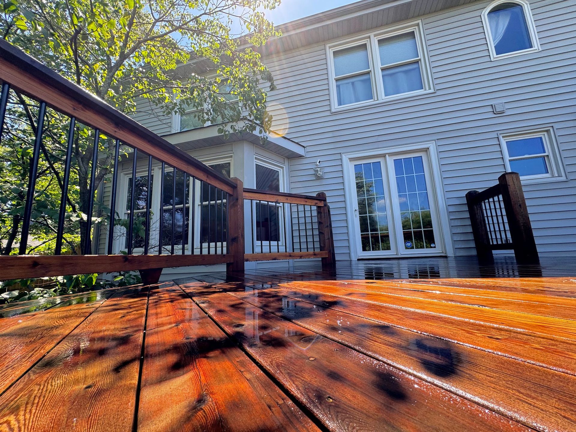Wooden deck with railing reflecting light, leading to a house with glass doors and blue siding.