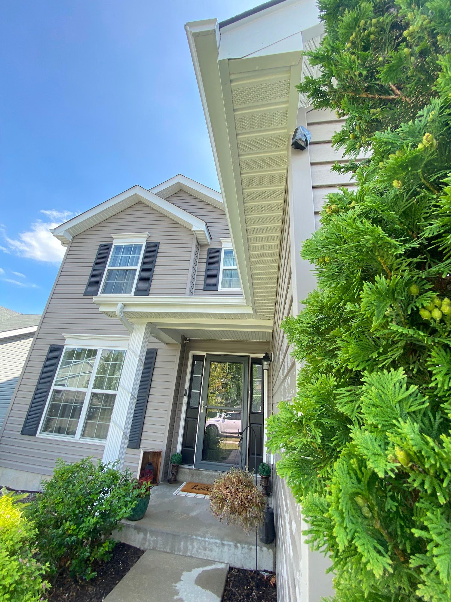 Tan two-story house with black shutters, white trim, and a small porch under a blue sky. Green bushes on the right.
