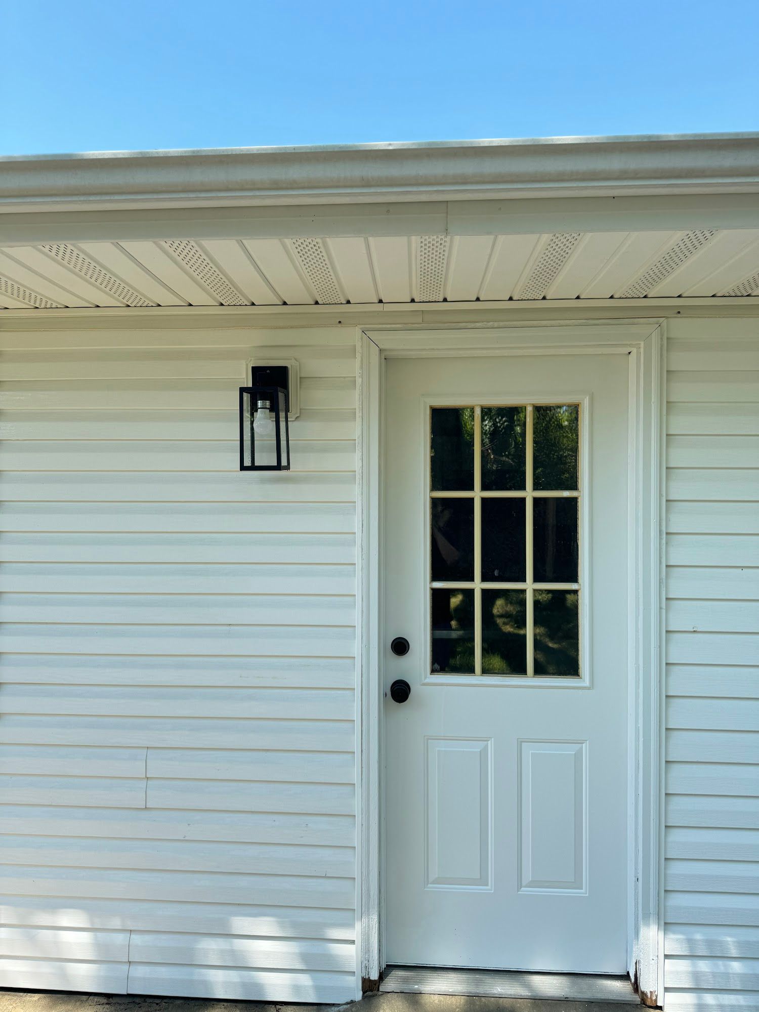 White exterior door with glass panes and black light fixture on white siding under blue sky.