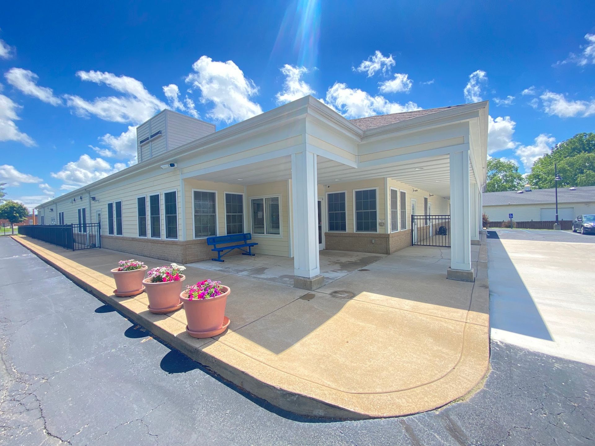 Exterior view of a light-colored building with a covered entrance, potted flowers, and a blue bench on a sunny day.