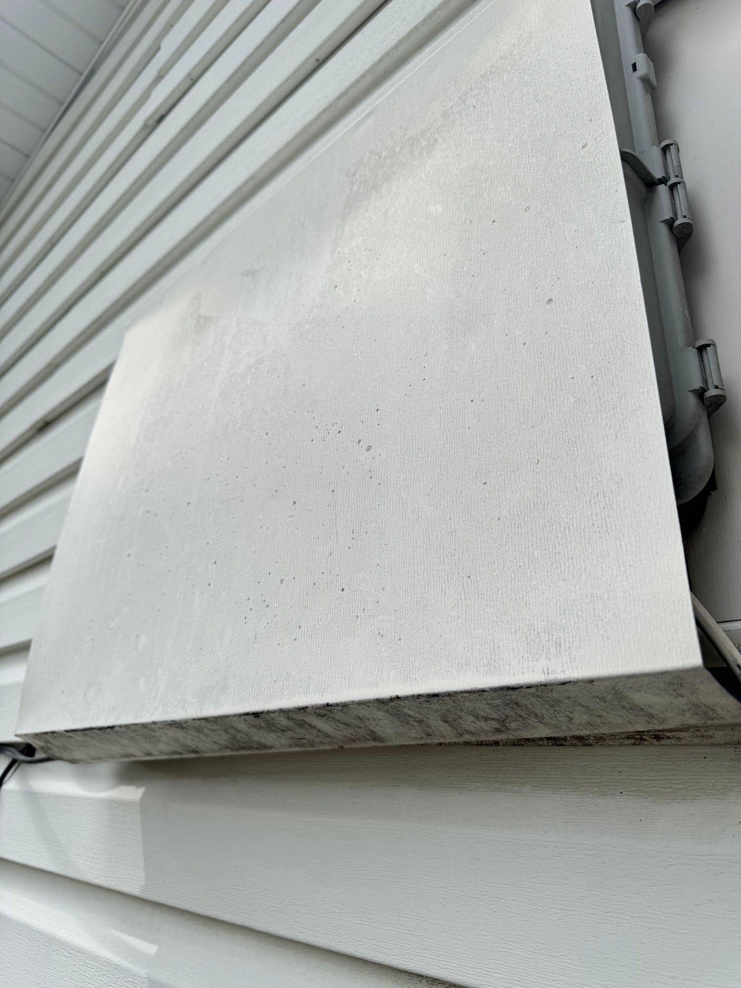 A rectangular concrete cover on a white-sided building with a pipe on the right.