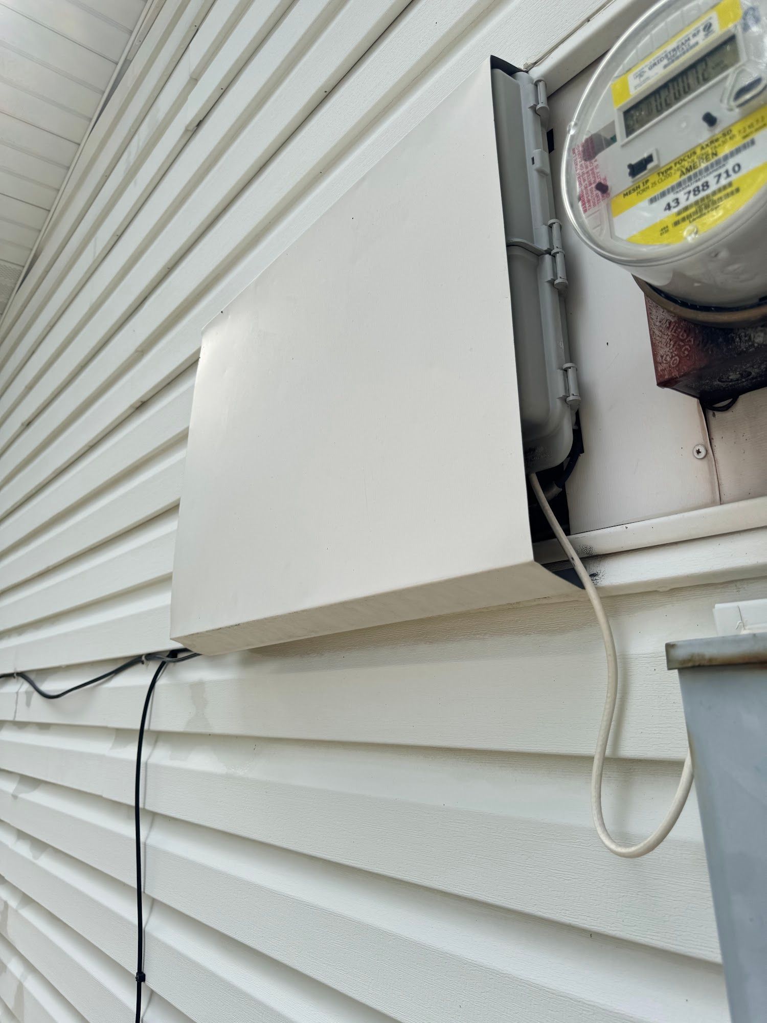 White electrical box mounted on a white-sided house next to a utility meter. A black and white wire are visible.