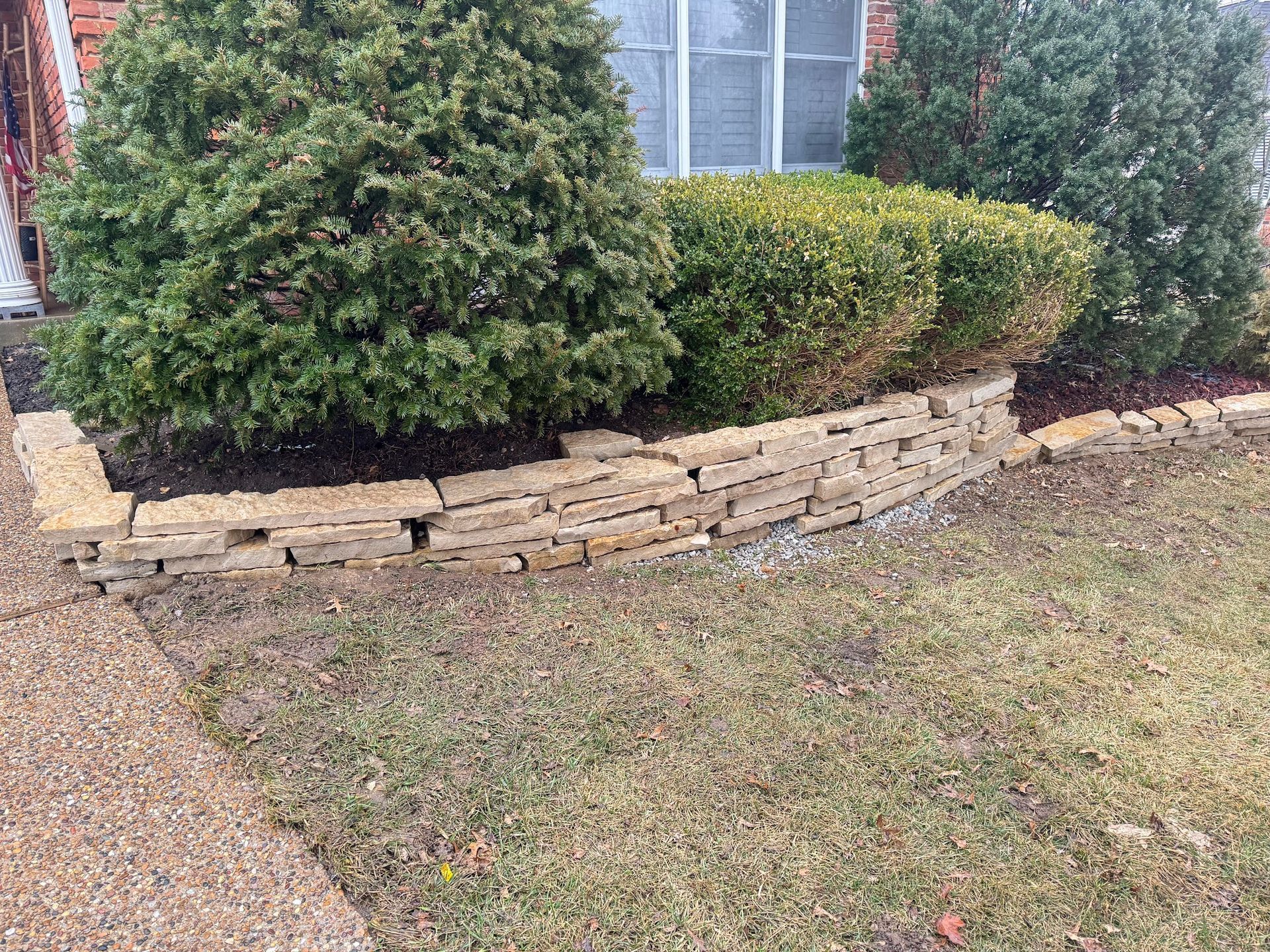 Low stone retaining wall with green bushes, next to a sidewalk and grass.