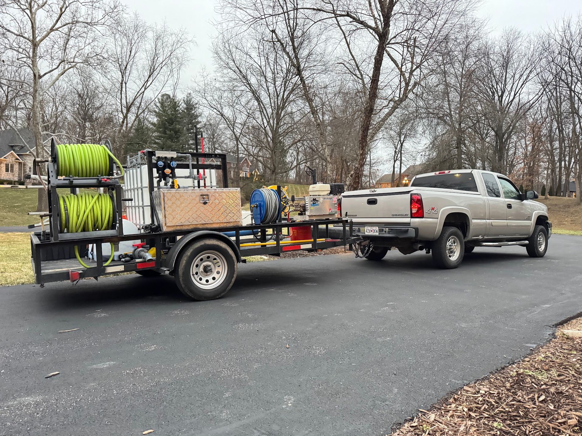 Truck towing a trailer with equipment, parked on a paved driveway in front of a residential area with bare trees.