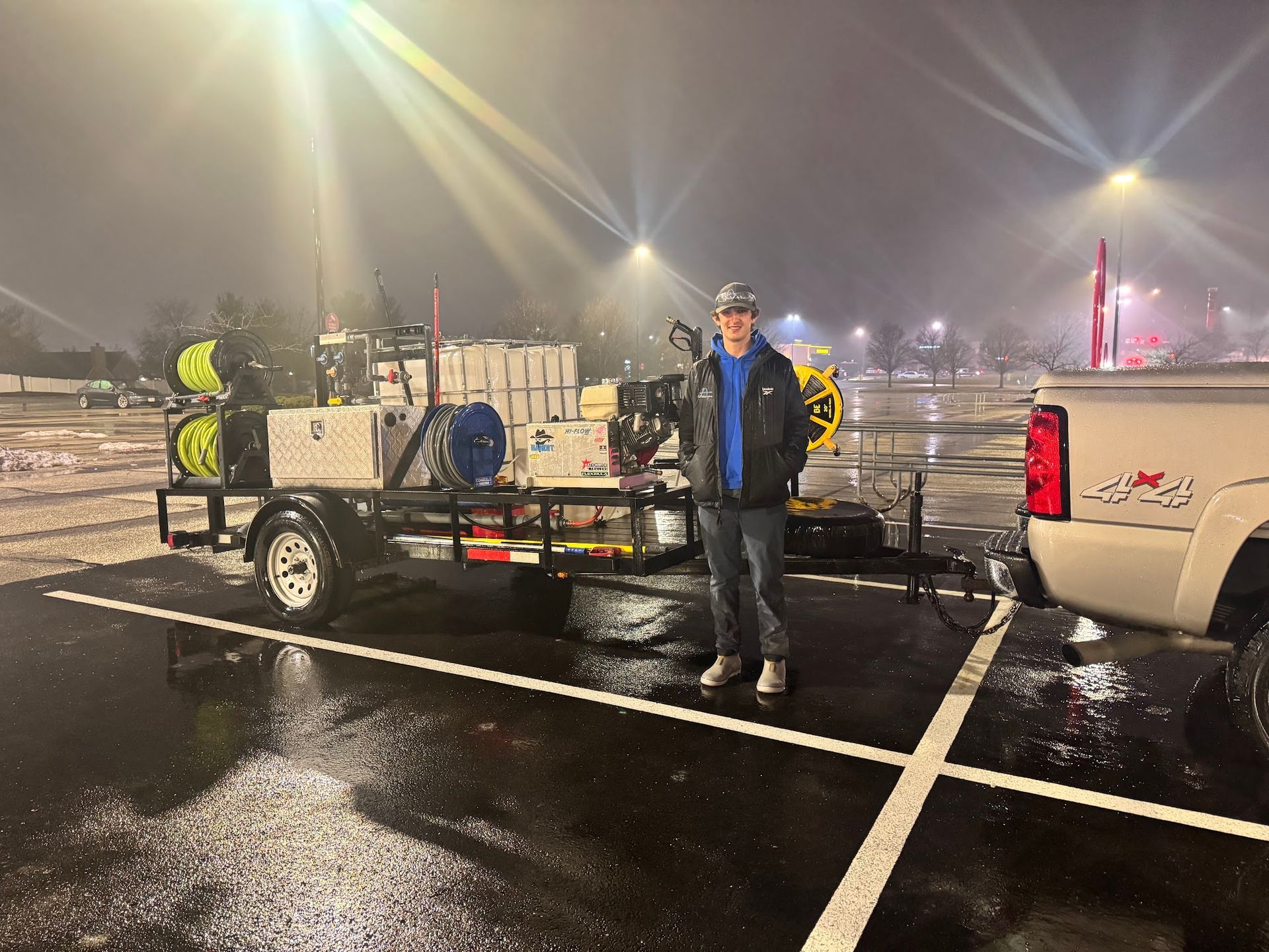 Man standing next to a trailer with equipment, parked in a wet parking lot at night.