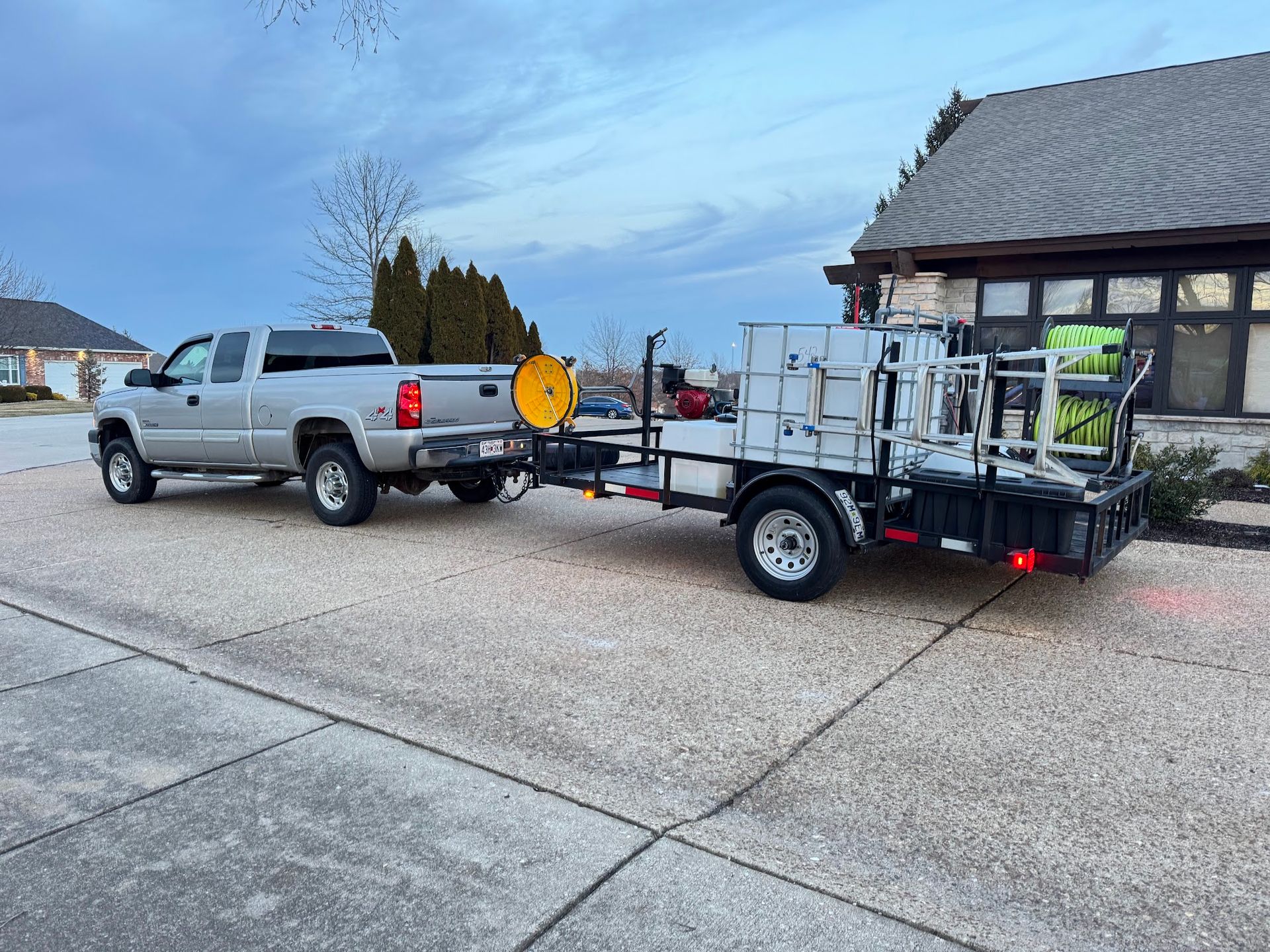 Silver truck towing a white trailer with equipment parked in front of a house on a cloudy day.