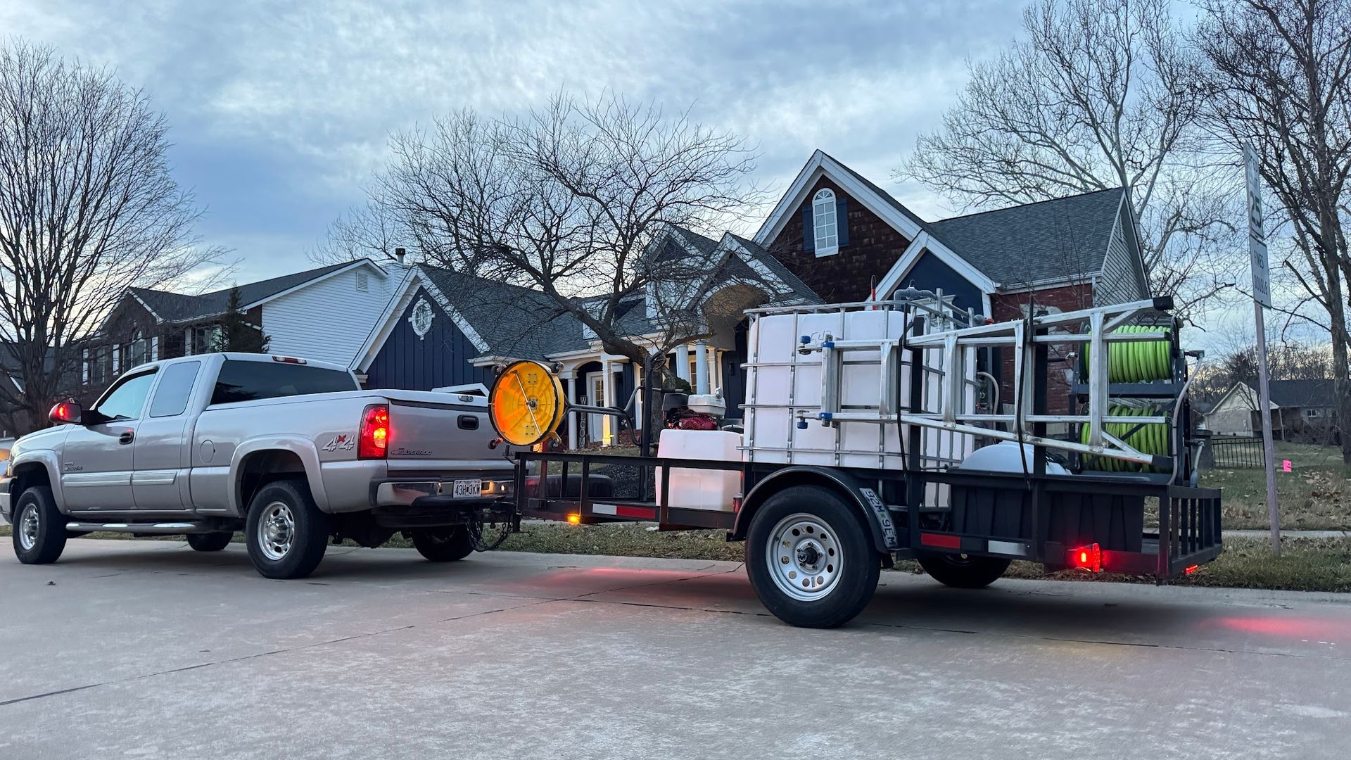 Silver truck towing a trailer with equipment, parked in front of houses on a cloudy day.