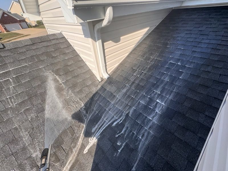 Washing a dark asphalt shingle roof with a high-pressure water stream near white siding and a gutter.