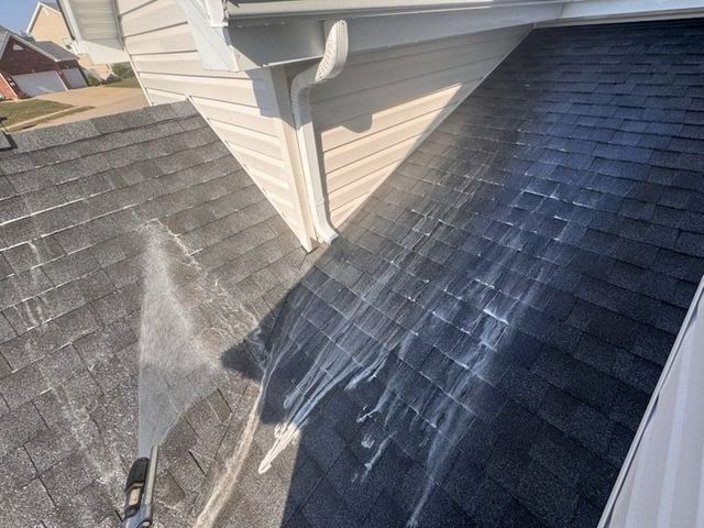 Washing a dark asphalt shingle roof with a high-pressure water stream near white siding and a gutter.