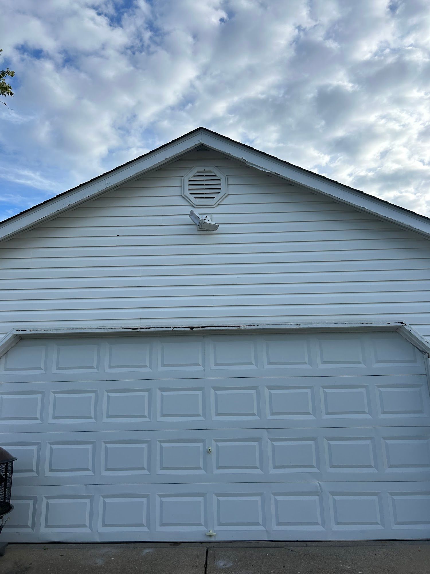 White garage with closed door, gable roof, and circular vent against a cloudy sky.