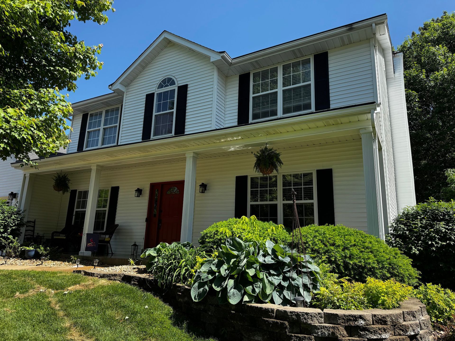 Two-story white house with black shutters, red door, and front porch. Green bushes and lawn.