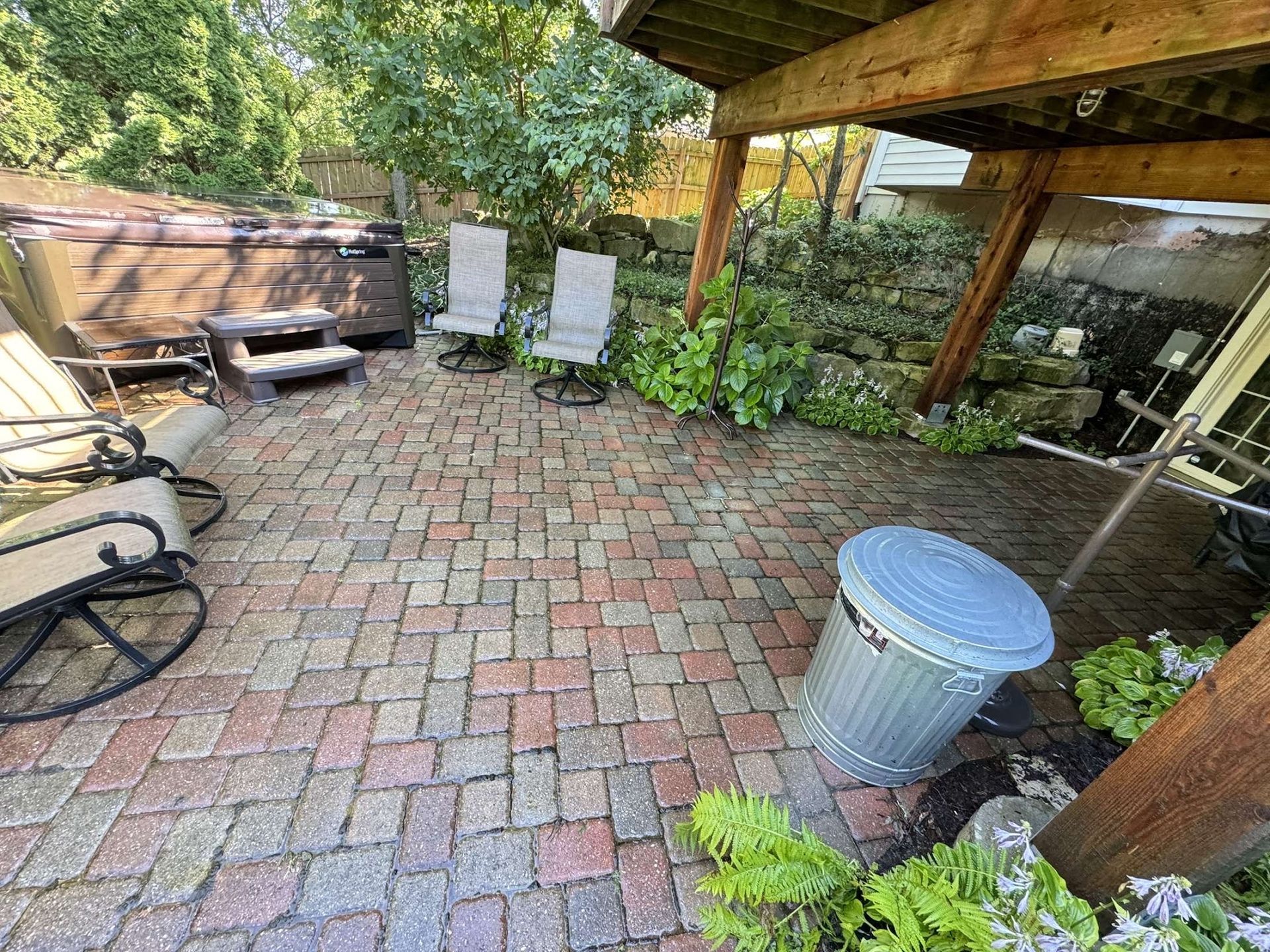Brick patio with outdoor seating, hot tub, and trash can under a wooden deck.
