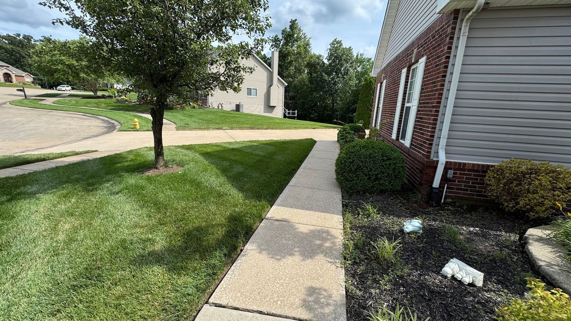 Sidewalk alongside a house with red brick accents and landscaping, leading to a street.