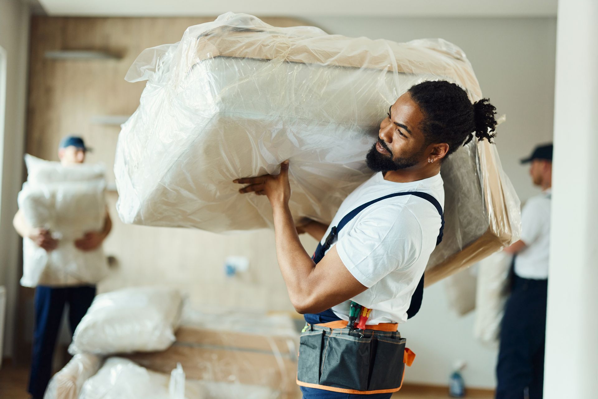 A black removalist, wearing a dark overall, carrying cream furniture in a new apartment.