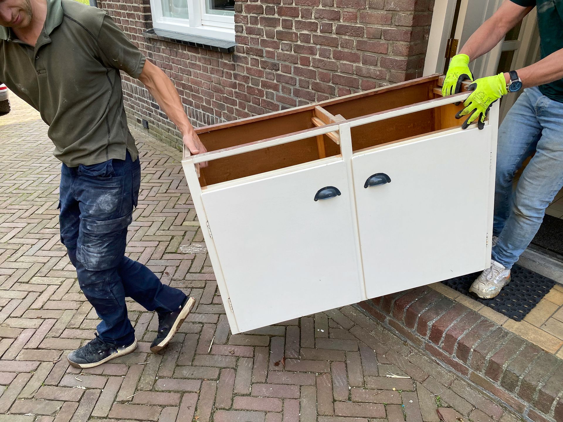 Two people carrying a white cabinet outside a brick building.