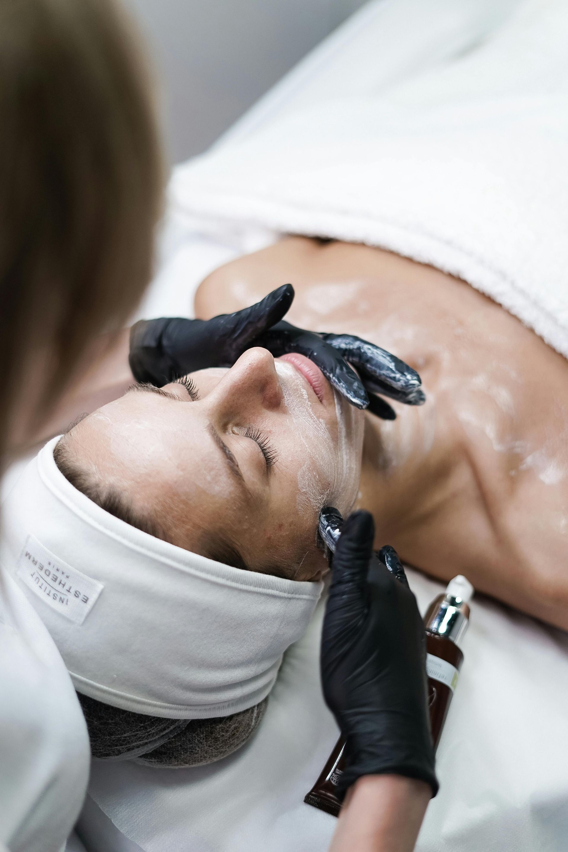 Facial treatment: Person receiving facial, lying down, with black-gloved hands applying product; salon setting.