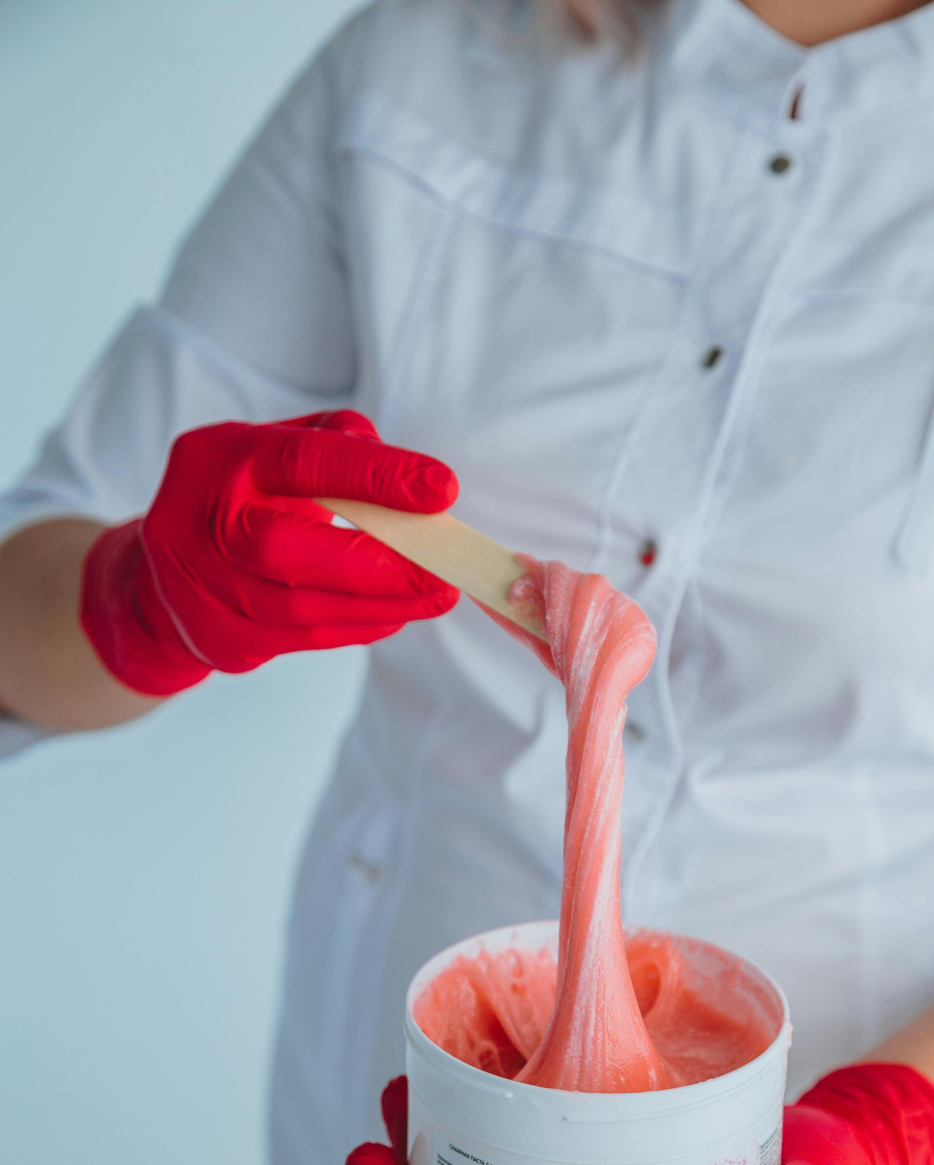 A person wearing red gloves and a white coat holds a spatula with pink waxing product.