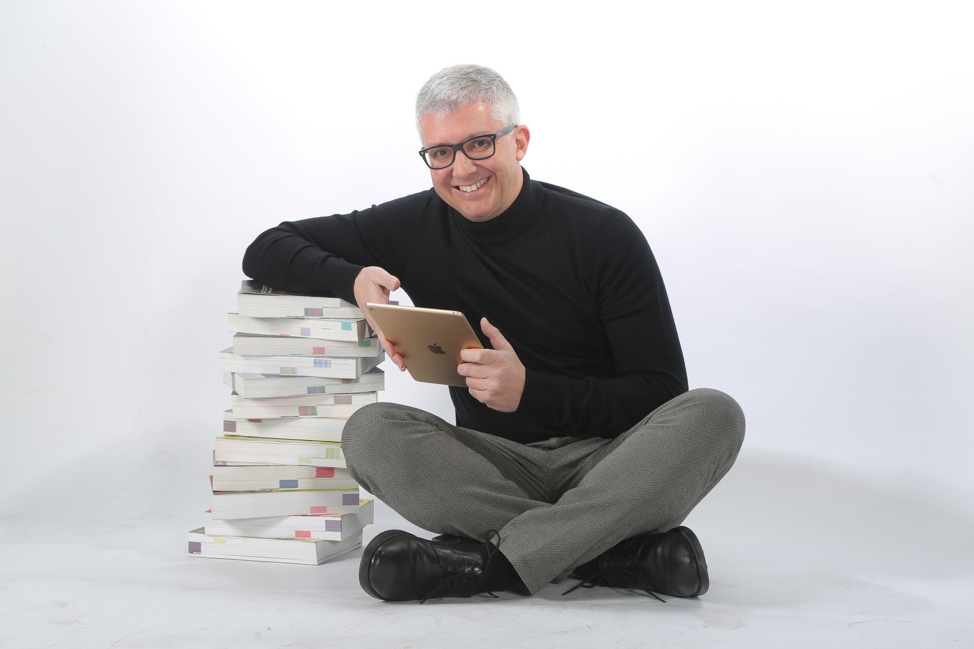 Un uomo è seduto sul pavimento con in mano un tablet accanto a una pila di libri.