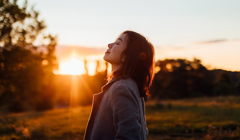 A woman is standing in a field at sunset with her eyes closed.