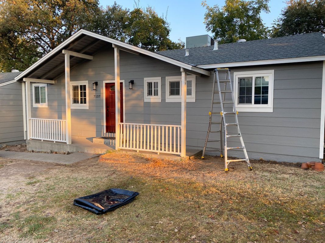 A gray house with a porch and a ladder in front of it.
