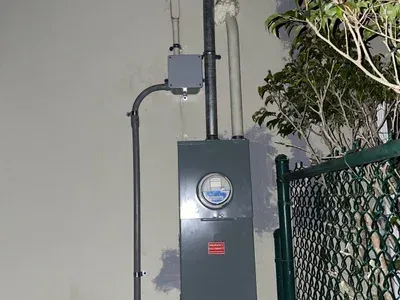 There is a meter on the side of a building next to a chain link fence.