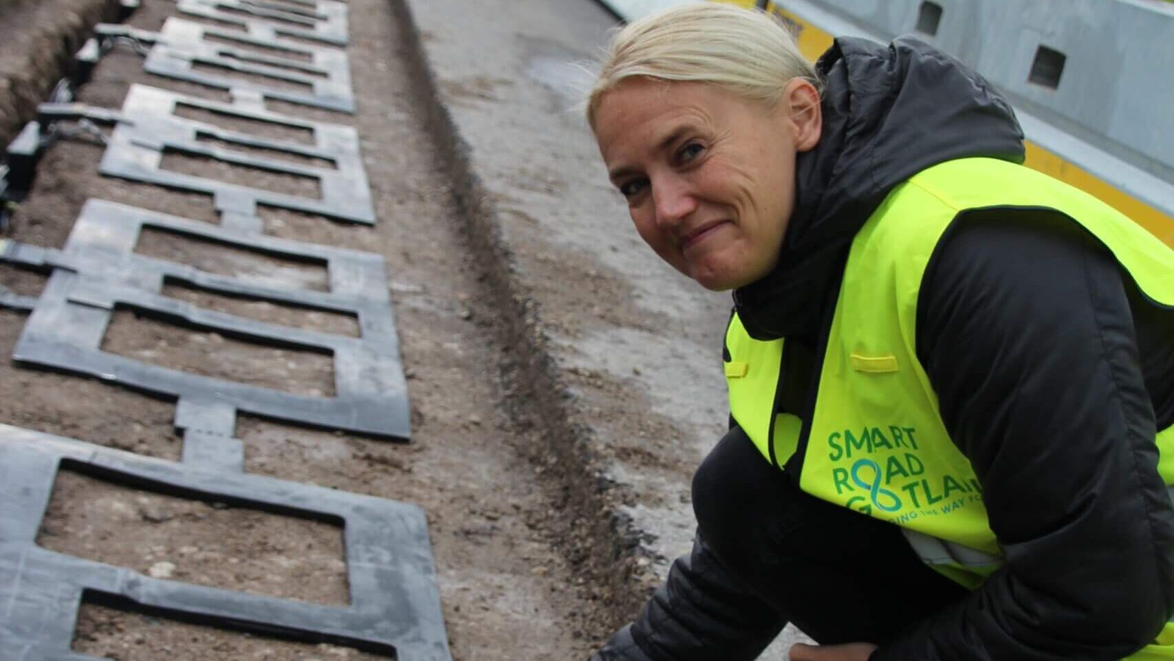 A smiling woman wearing a yellow safety vest squats beside a construction road of Smart Road.