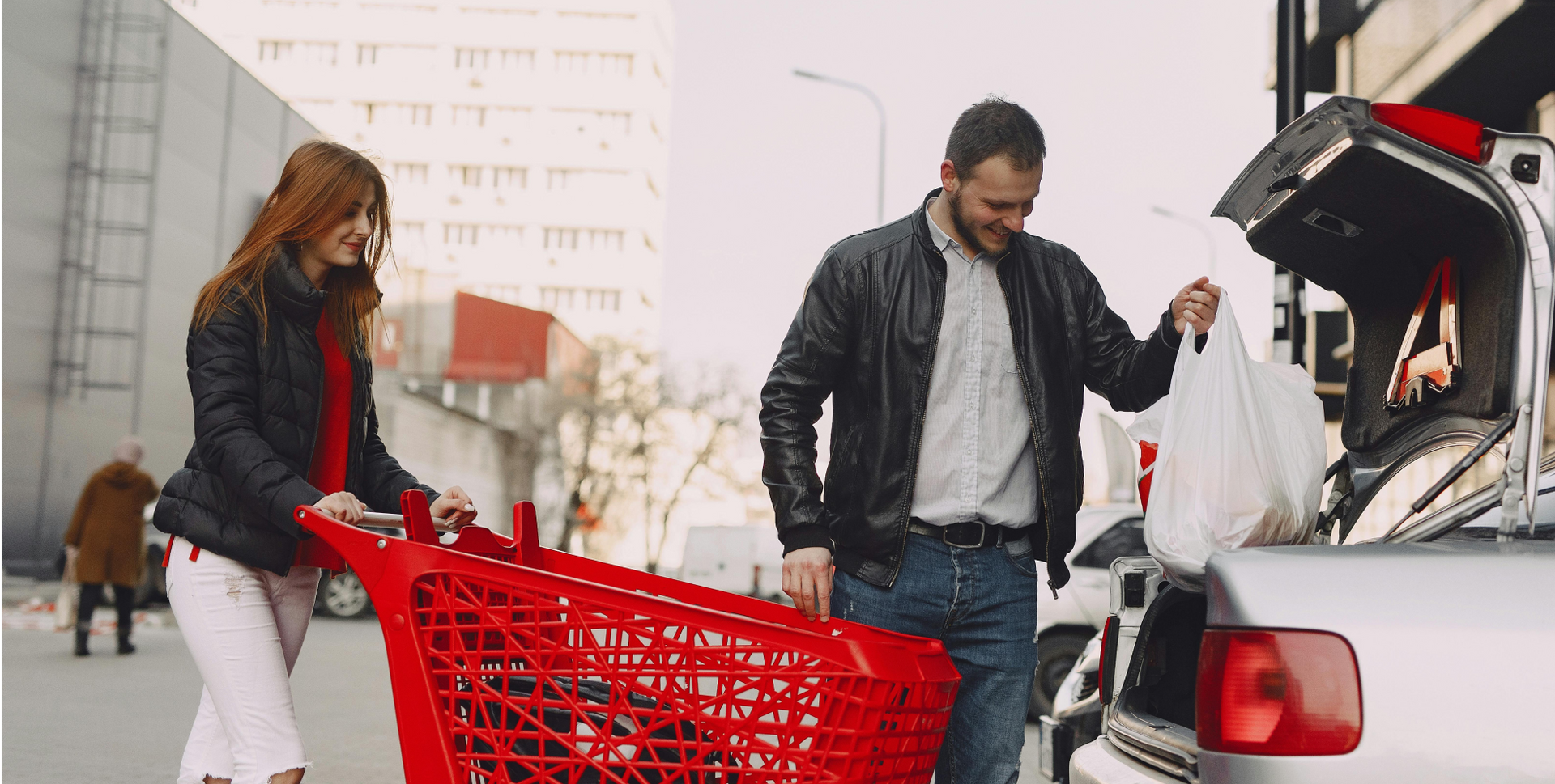 A man and a woman are pushing a red shopping cart near their car and putting the purchased goods in the trunk.
