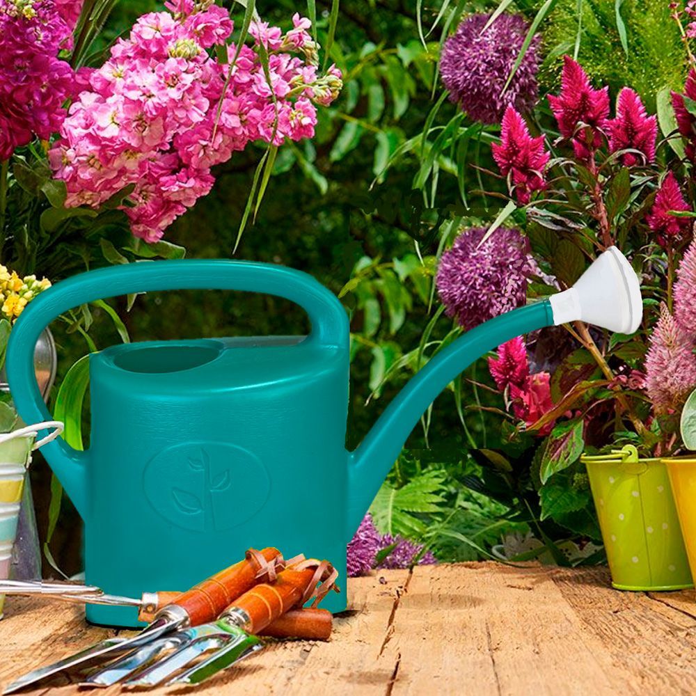 A watering can and gardening tools are on a wooden table in front of flowers.
