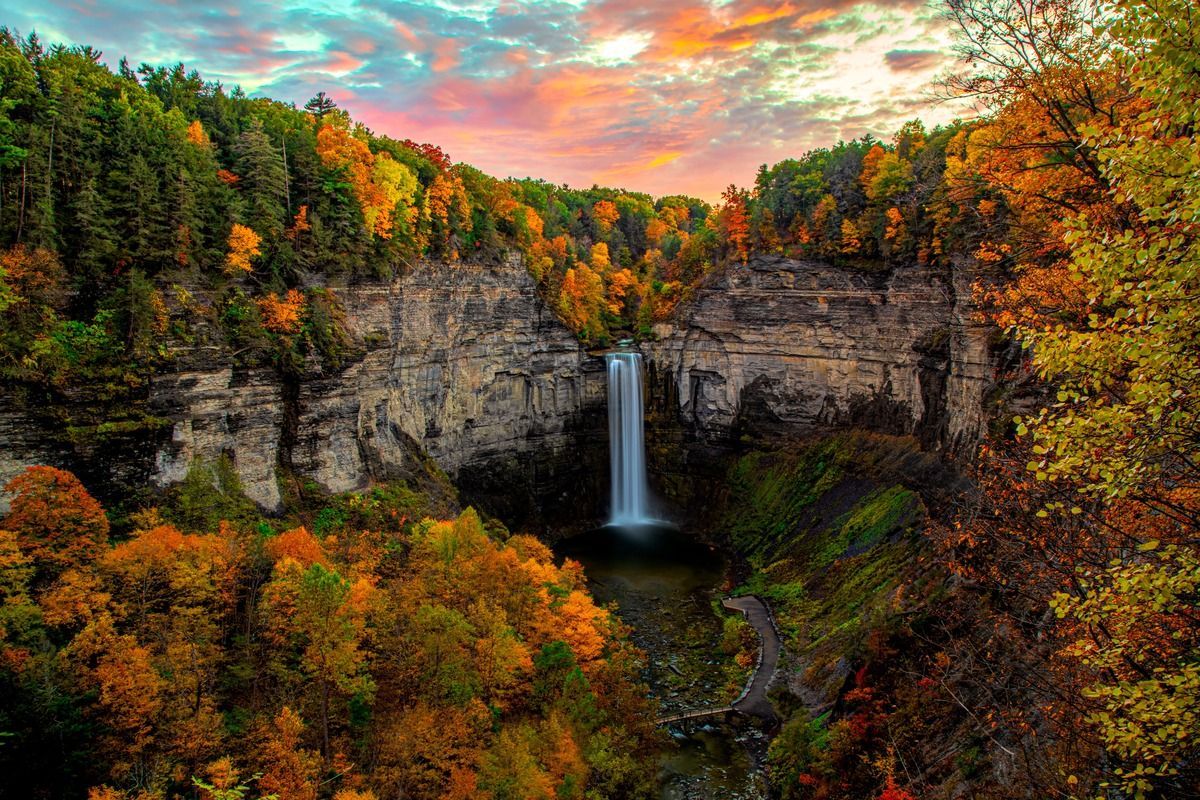 Waterfall cascading down a rocky cliff, surrounded by autumn foliage under a colorful sunset.