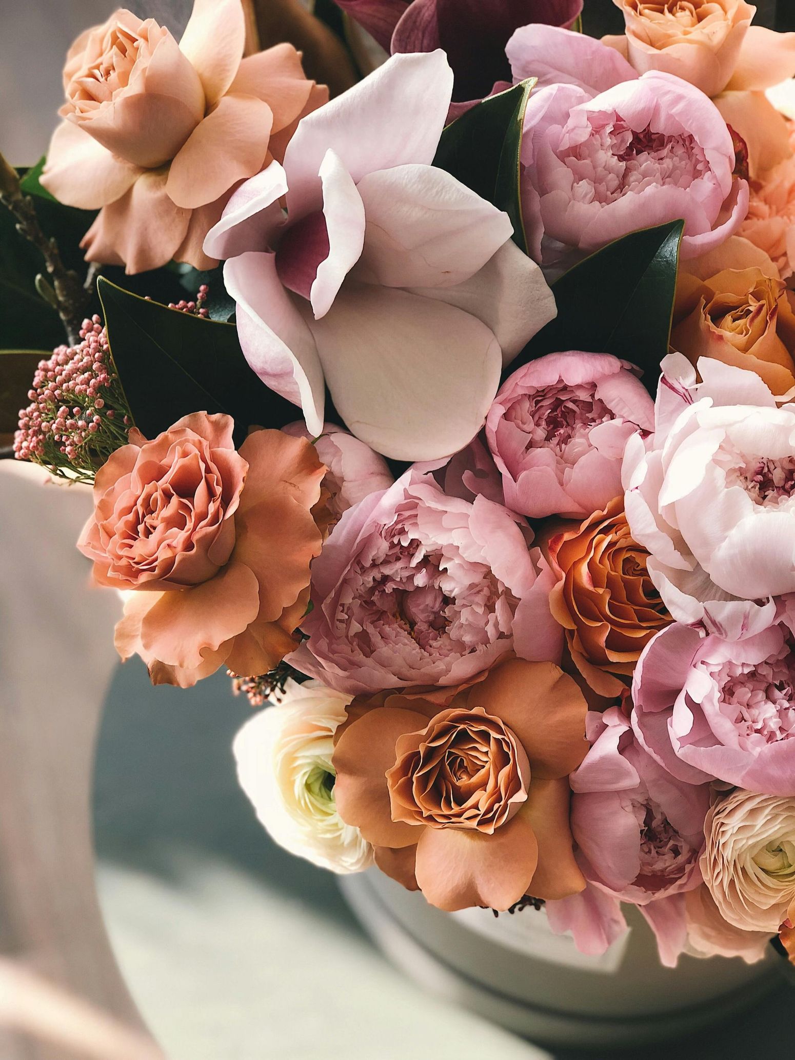Close-up of a bouquet with peach, pink, and white peonies, orchids, and other flowers in a light-colored vase.