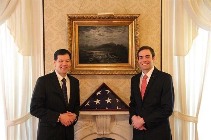 Two men in suits stand near a fireplace with a folded American flag and a landscape painting.