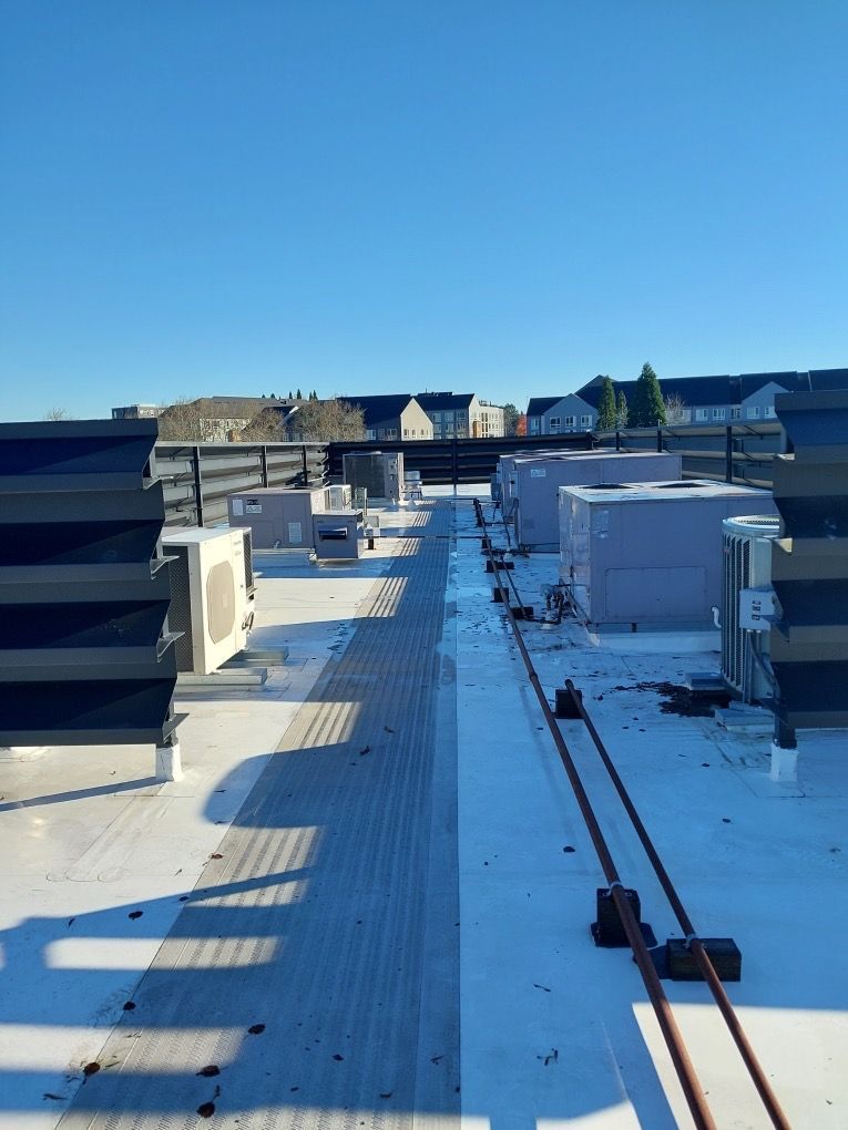 A view across a flat commercial rooftop showing multiple HVAC units, a walkway, and utility piping under a clear blue sky.