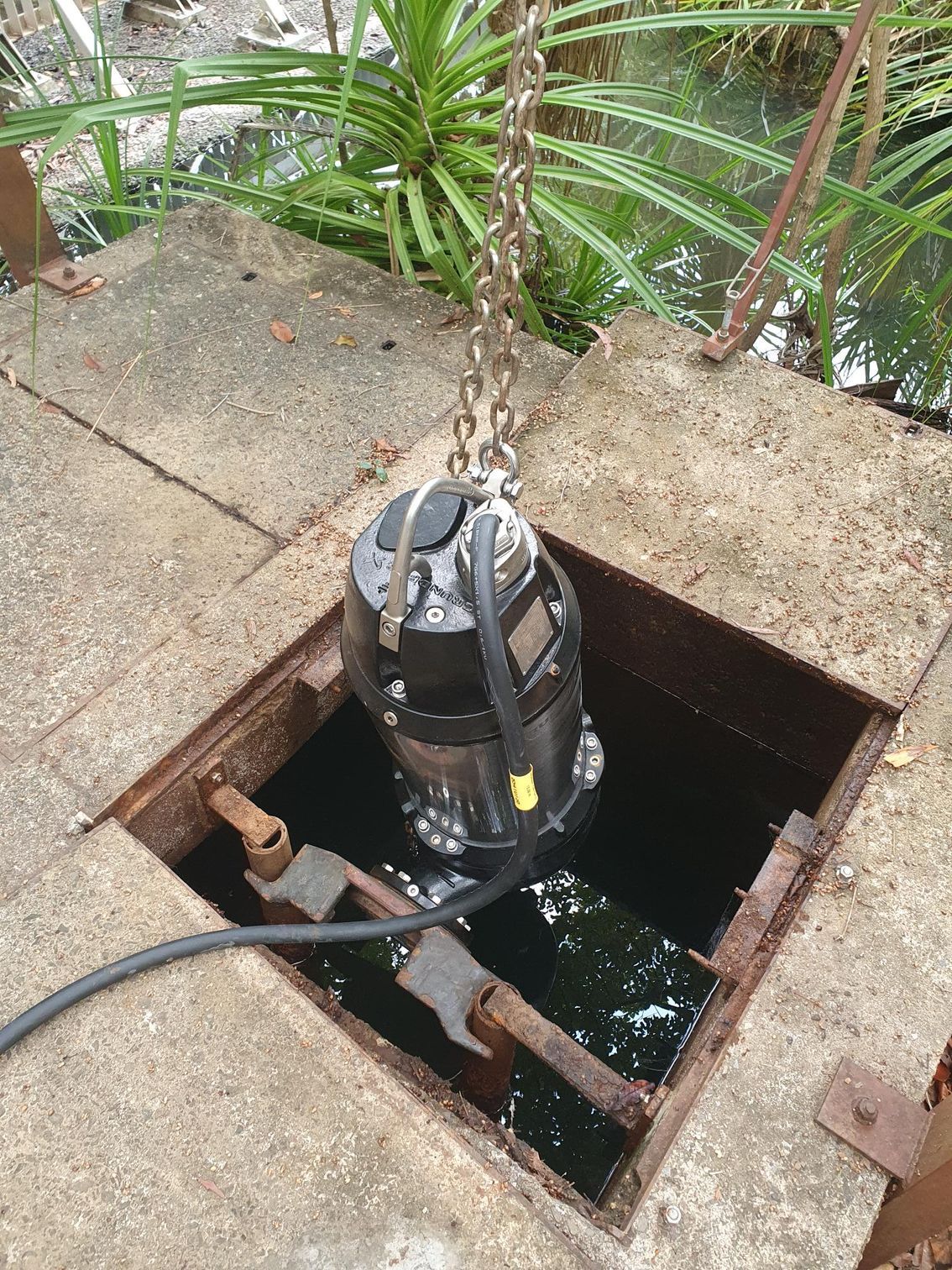 A pump is hanging from a chain in a hole in the ground.