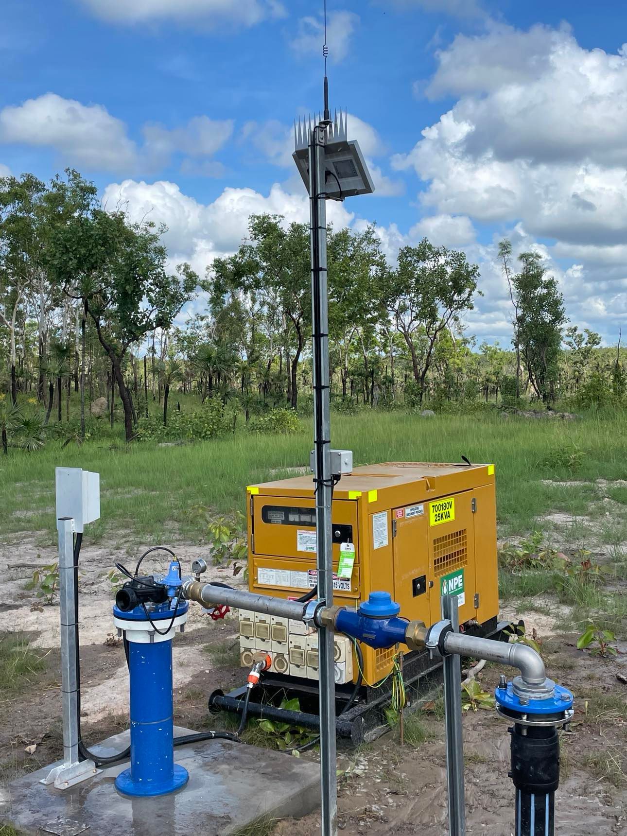 A yellow generator is sitting in the middle of a field next to a blue pipe.