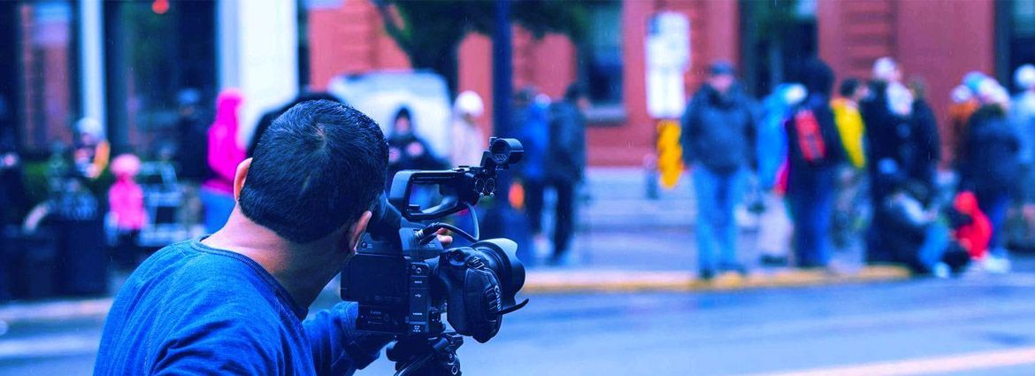 A videographer seen from behind his camera which is pointing across the street at a group of people in the blurry background