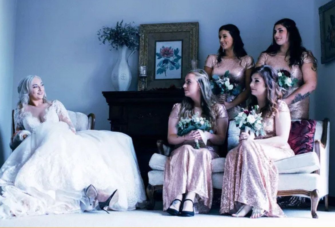 A bride in a beautiful wedding dress  chatting to her four bridesmaids sitting on the right, holding flowers.