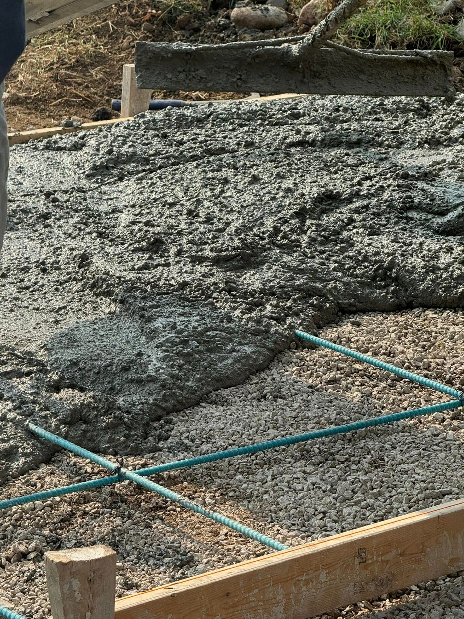 Pouring concrete onto a gravel base, with blue tubes, wooden forms, and a person's leg visible.