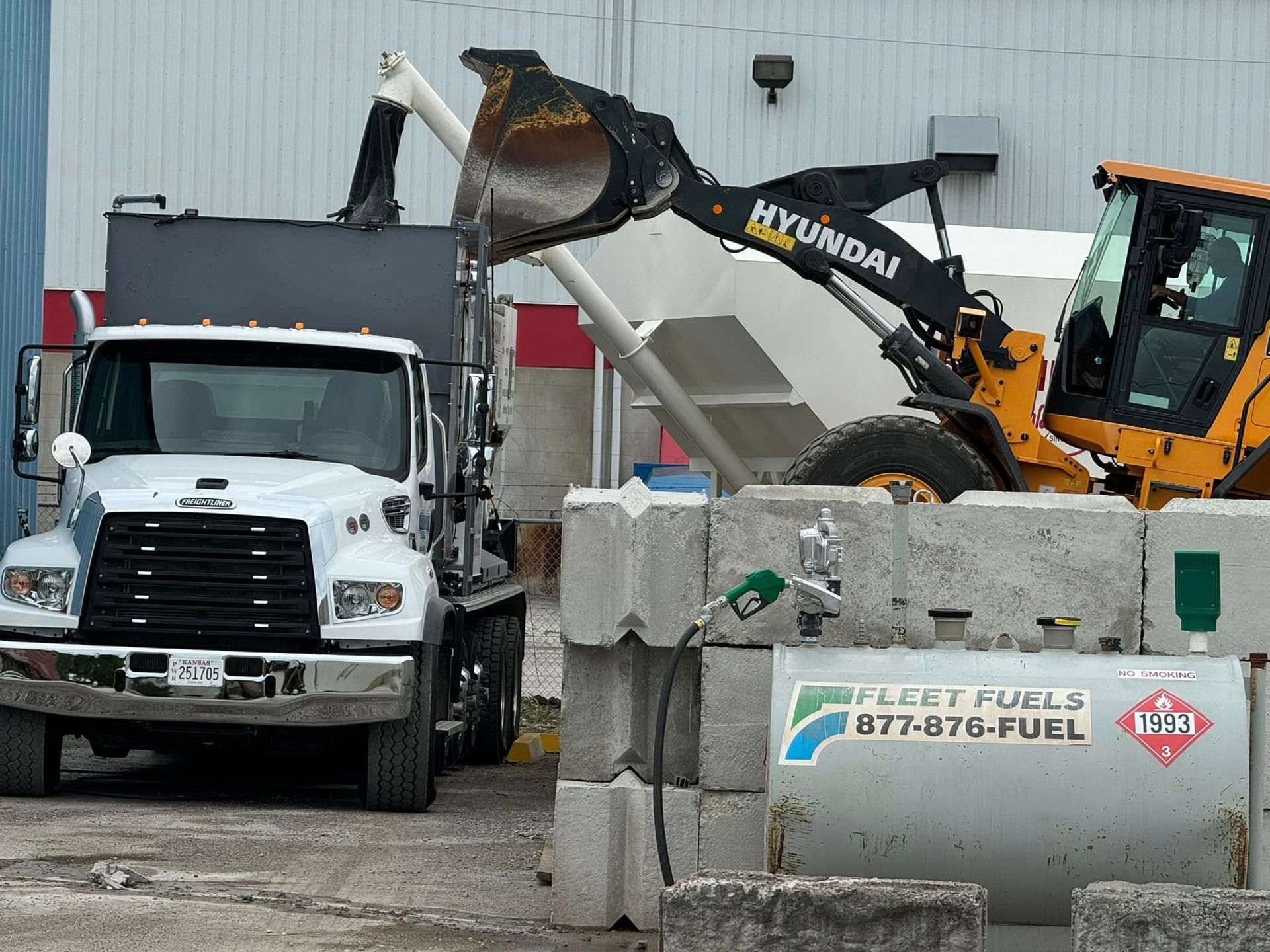 A front loader dumps material into the bed of a white dump truck parked near a fuel tank and concrete barrier blocks.