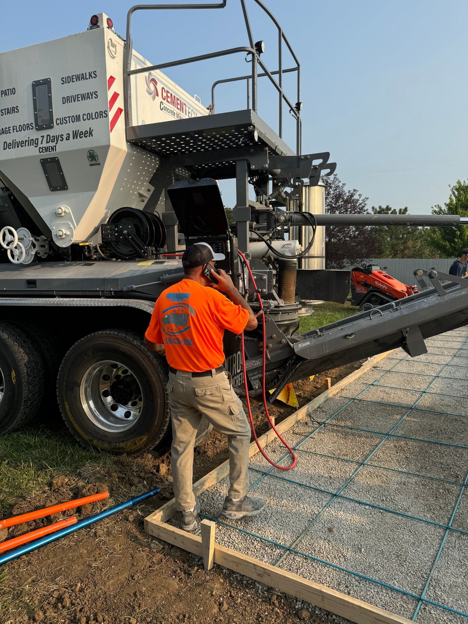 A worker in an orange shirt talks on a phone while operating a concrete mixing truck beside a prepared gravel site.