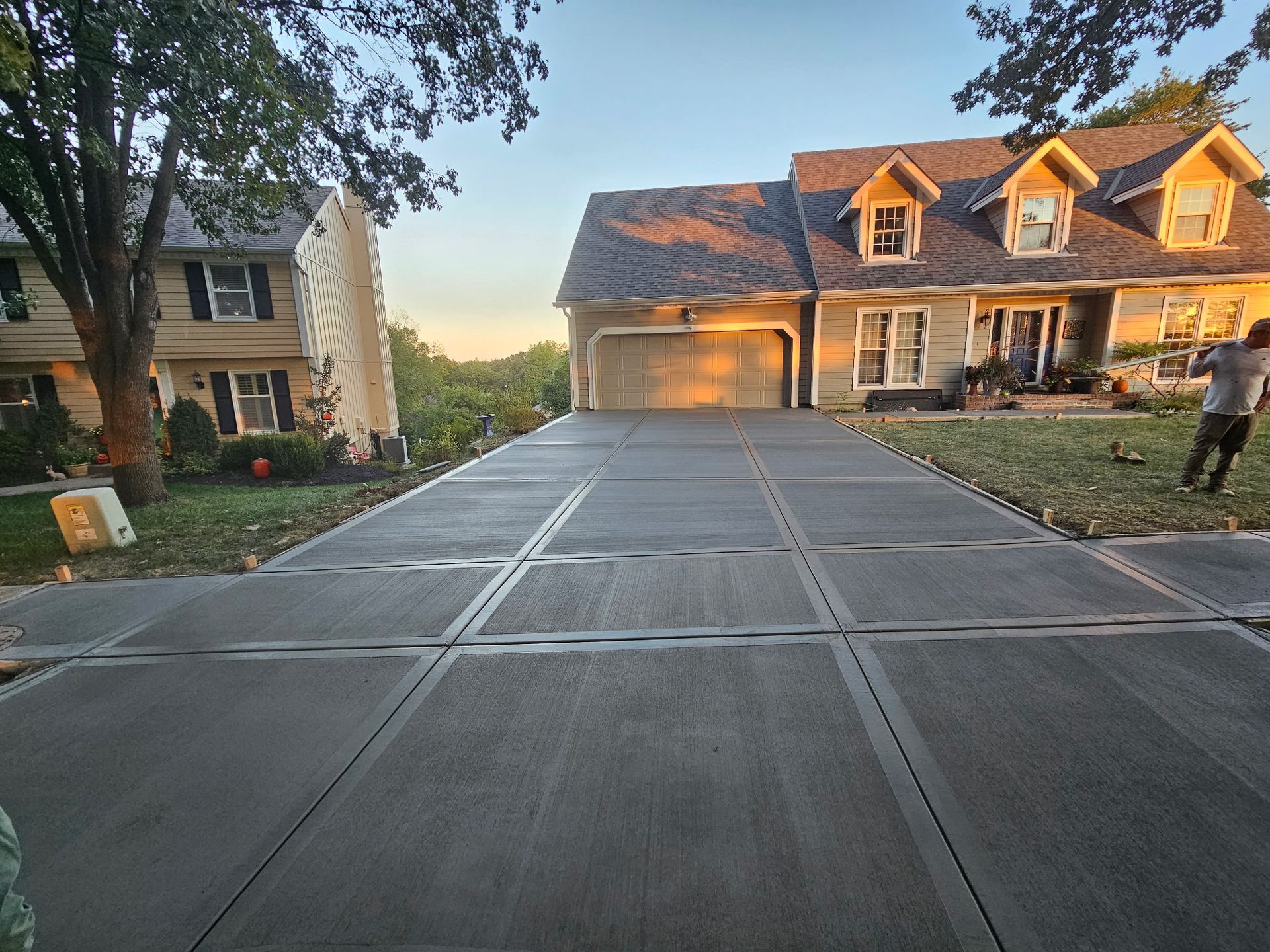 A wide view of a recently poured concrete driveway leading to a suburban home with a person standing on the lawn.